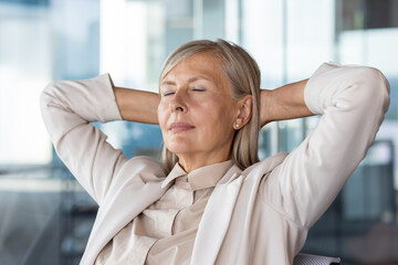 Close-up photo of a senior gray-haired business woman in a suit sitting in a chair in the office and resting, hands behind her head and eyes closed. - Powered by Adobe