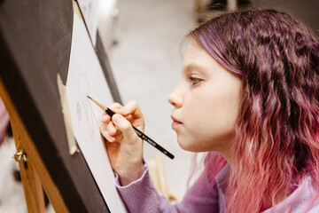 Girl 11 years old craftswoman are painting on canvas in studio standing in front of easel. Portrait of a girl painting during an art class.