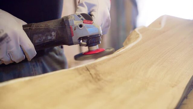 Craftsman in protective gloves using electric grinding machine for sharpening wood plank angles in workshop, sharp rotating disc of grinding machine removing excessive wood making smooth wood bends