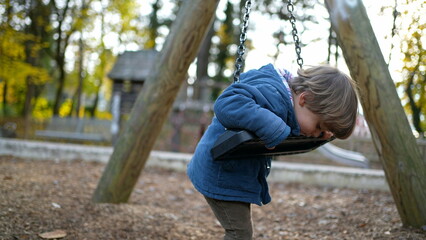 Carefree little boy leaning on park swing during autumn day wearing blue jacket, child twisting and turning into swing playing by himself during fall season outdoors