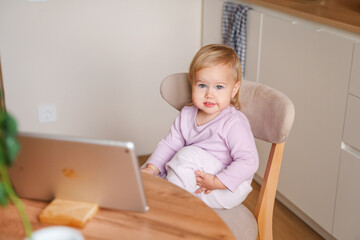 Cute baby girl toddler child sits in the kitchen in front of a tablet and watches cartoons or communicates with tom videos with relatives. Saint Petersburg, Russia - 19 Nov 2023