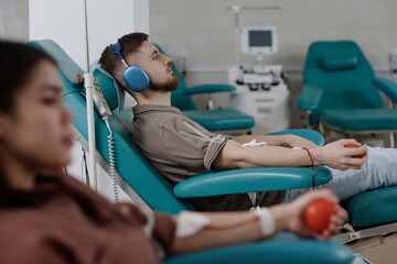 Young man and woman sitting in medical chairs during blood transfusion, focus on man in headphones