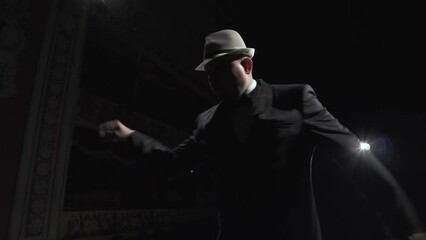 Man in suit puts on hat and starts dancing. Dancer on dark stage of concert hall in the backlight of soffit.