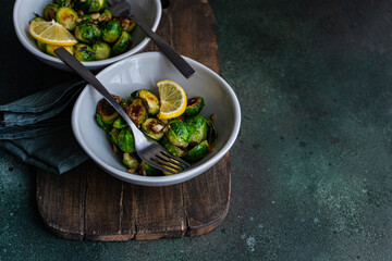 From above of bowl of grilled Brussels sprouts seasoned with garlic and spices, garnished with a slice of lemon, served on a wooden board with a fork