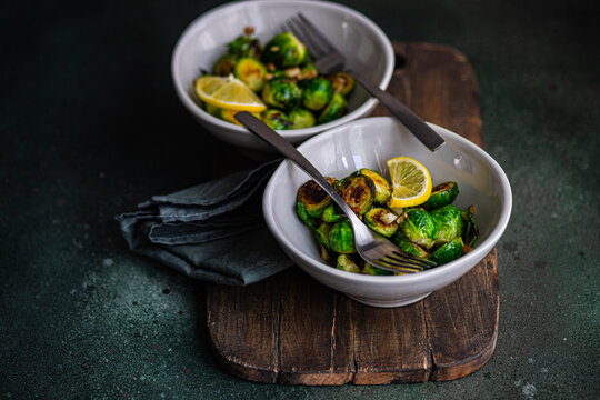 From above of bowl of grilled Brussels sprouts seasoned with garlic and spices, garnished with a slice of lemon, served on a wooden board with a fork