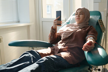 Muslim woman relaxing on medical chair and scrolling smartphone during blood transfusion in hospital - obrazy, fototapety, plakaty