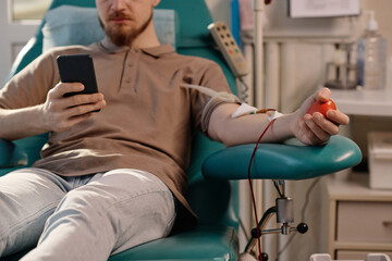 Male blood donor sitting in medical chair and browsing his smartphone during transfusion process
