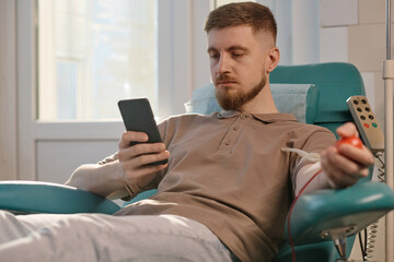 Young male donor browsing his smartphone sitting in medical chair during blood transfusion in clinic