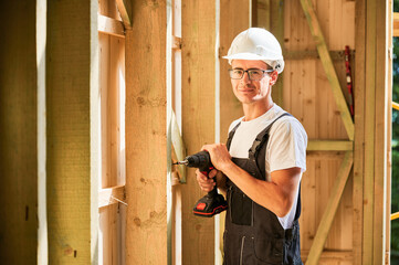 Carpenter constructing wooden framed house. Man worker in glasses working with screwdriver, wearing work overalls and helmet. Concept of modern eco-friendly construction.