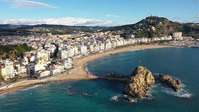 Scenic drone view of coastal Catalan town of Blanes overlooking white residential buildings and Sa Palomera Rock on background of San Juan hill with ancient castle on sunny day, Barcelona, Spain 