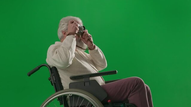 Portrait of aged disabled man on chroma key green screen. Close up senior man in wheelchair taking pictures on old film camera.