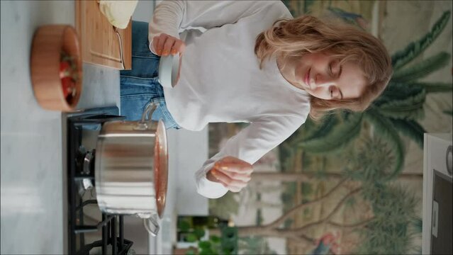 Vertical video. Girl salts soup. A young woman prepares homemade healthy food in her home kitchen. Adding seasonings to a dish. Stirs  hot soup with a spoon