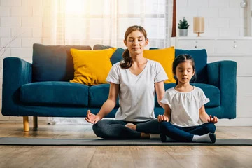 Tuinposter A beautiful young woman and her charming daughter share smiles during their family yoga session at home emphasizing mindfulness and meditation in lotus position creating happiness and togetherness. © sorapop
