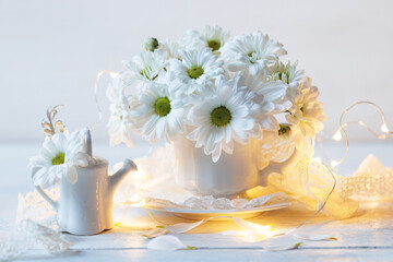 Chrysanthemum flowers in a white cup, watering can, light garland and lace ribbon on a white wooden table. Beautiful still life, postcard for the holiday.