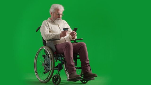 Portrait of aged man on chroma key green screen. Senior man in wheelchair inserts card number in smartphone, paying online.