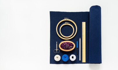 Supplies for hand embroidery in Sashiko style: blue canvas, white threads, pincushion, needles, pins, scissors, wooden hoops, tailor's pencil, ruler on white table before work. Flat lay, copy space