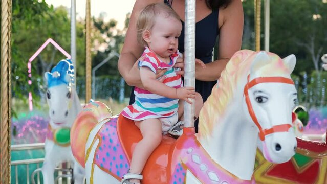 Young mother sitting her baby on a vintage horse carousel in funfair or an amusement park in the evening, slow motion. Happy childhood and memorable moments concept