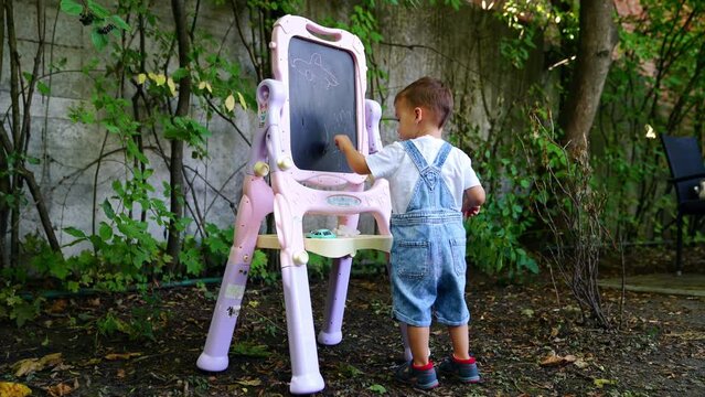 Two year old toddler wearing jeans romper draws with the chalk on the blackboard. Future artist concept.