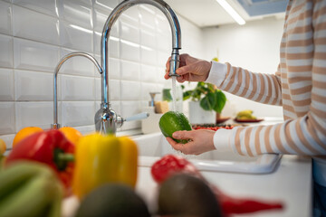 Colourful vegetables for healthy eating on cook table. Hands holding wash green pepper under water tap on light home kitchen. Clean paprika for prepare tasty vegan salad. Vegetarian raw organic food