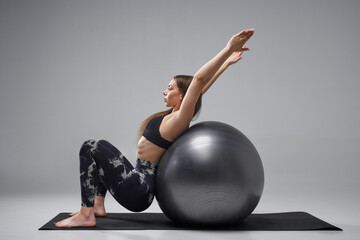 Flexible young woman doing yoga exercise on fit ball. Side view of beautiful slim female using silver pilates ball, while exercising in studio, isolated on grey background. Fitness, pilates concept.