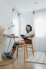 Woman with prosthetic leg sitting in chair in living room at home. Relaxed, happy woman at home living room sitting on the chair, using tablet