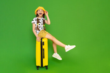 A young girl with a yellow suitcase is going on a trip. A happy teenage girl in a summer hat and shorts is sitting on her luggage and smiling broadly. Green isolated background.