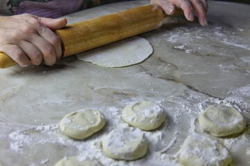 on the kitchen table, women's hands roll out dough with a rolling pin. several round pieces of dough and a prosky piece for stuffing it with meat are visible.