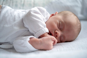 Close Up Photography of Peaceful newborn baby sleeping on its left side, radiating innocence and calm. Sleeping peacefully on its side, wearing white pajama on white bed. White bedsheet.
