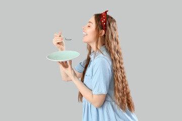 Beautiful young woman with empty plate and spoon on grey background