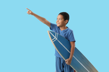Cute little African-American boy with surfboard pointing somewhere on blue background