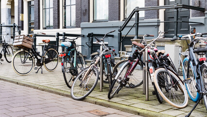 Fototapeta na wymiar Old recreational bikes carelessly parked along a street near a city building in Amsterdam. A bicycle parking area on a city street. Public bicycle transport in a parking lot. An urban cityscape.