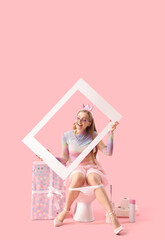 Young woman with frame sitting on toilet bowl against pink background
