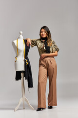 happy trendy asian stylist posing near mannequin with fabric and measuring tape on grey backdrop
