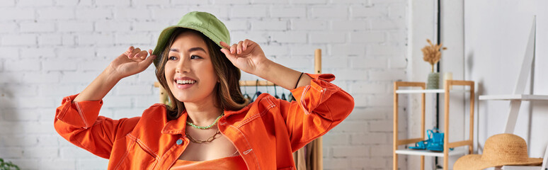 happy asian stylist in orange attire wearing panama hat in clothes atelier, fashion business, banner