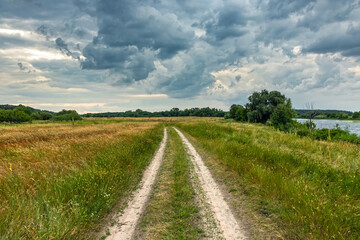 Landscape of a picturesque river valley with a country road in a meadow and trees on a cloudy summer day