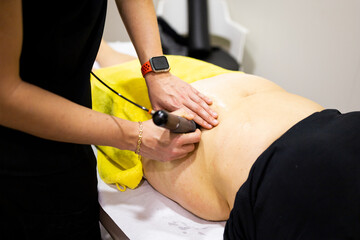 An unrecognizable physiotherapist uses shock wave therapy on a woman's abdomen in a modern clinic....