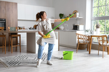 Pretty young woman having fun with mop in modern kitchen