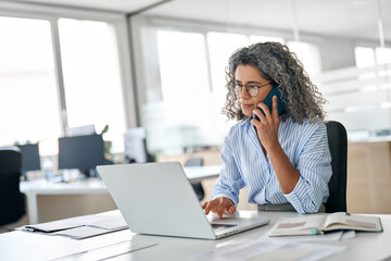 Busy mature middle aged business woman, senior professional lady executive manager talking on the phone making business call on cellphone sitting at work desk in office using laptop computer.