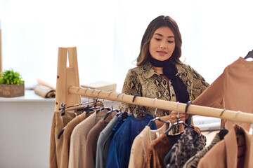 ambitious asian stylist standing near trendy casual clothes on rack in modern fashion studio