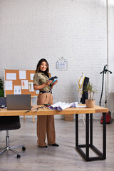 happy asian designer with color swatches near laptop and sewing patterns in modern fashion atelier