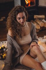 Young beautiful relaxed attractive woman with curly hair at home, wearing casual soft clothes. Winter, countryside house, fireplace. Cozy home holiday atmosphere, country interior, warm light