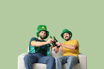 Young men in leprechaun hats with green beards holding bottles of beer and sitting on sofa against...