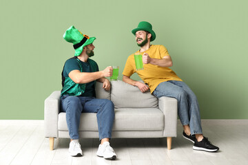 Young men in leprechaun hats with green beards holding glasses of beer and sitting on sofa near...