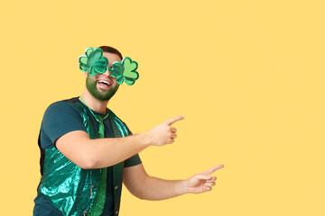 Young man in decorative glasses in shape of clover with green beard pointing at something on yellow...