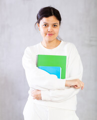 Fototapeta premium Portrait of smiling young Asian woman, tutor or college student, standing on gray studio background with workbooks in hands