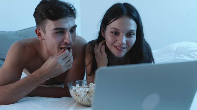 A cheerful young man and woman lie in bed at night, watching a movie on a laptop and sharing popcorn, enjoying quality time together.