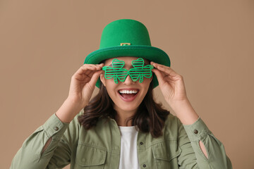Happy young woman in leprechaun hat and stylish decorative glasses in shape of clover on brown...