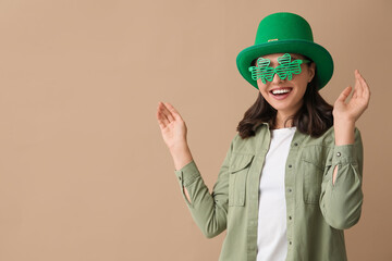 Happy young woman in leprechaun hat and stylish decorative glasses in shape of clover on brown...