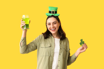 Happy young woman in leprechaun hat with glass of beer and clover on yellow background. St....