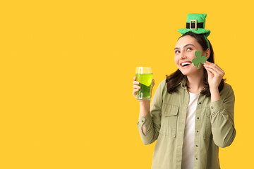 Happy young woman in leprechaun hat with glass of beer and clover on yellow background. St....